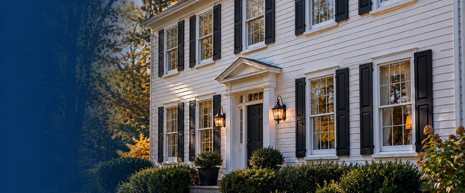 A two-story wood-sided colonial with tall original windows fitted with slim white aluminum storm windows, the old glass panes visible behind the outer layer, autumn leaves on the lawn